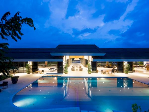 A modern luxury building with large glass windows is illuminated at dusk, featuring an inviting outdoor swimming pool in the foreground.