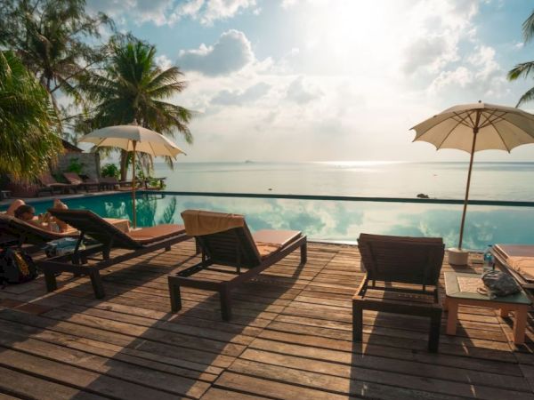 Loungers and umbrellas on a wooden deck overlooking a serene swimming pool with a stunning ocean view in the background, under a partly cloudy sky.