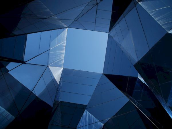 The image shows a geometric, abstract view of a building's glass exterior, looking up through a partially open, angular skylight against a blue sky.