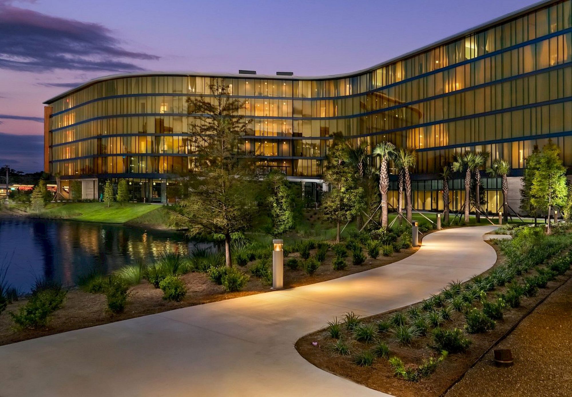 A modern, multi-story building with large glass windows, surrounded by a well-lit path, trees, and landscaping, near a body of water at twilight.