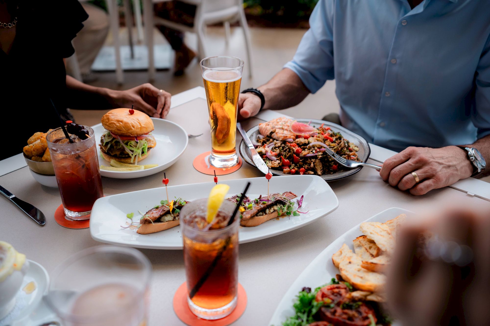 People enjoying a meal with drinks, including a burger, salad, and appetizers at a dining table.