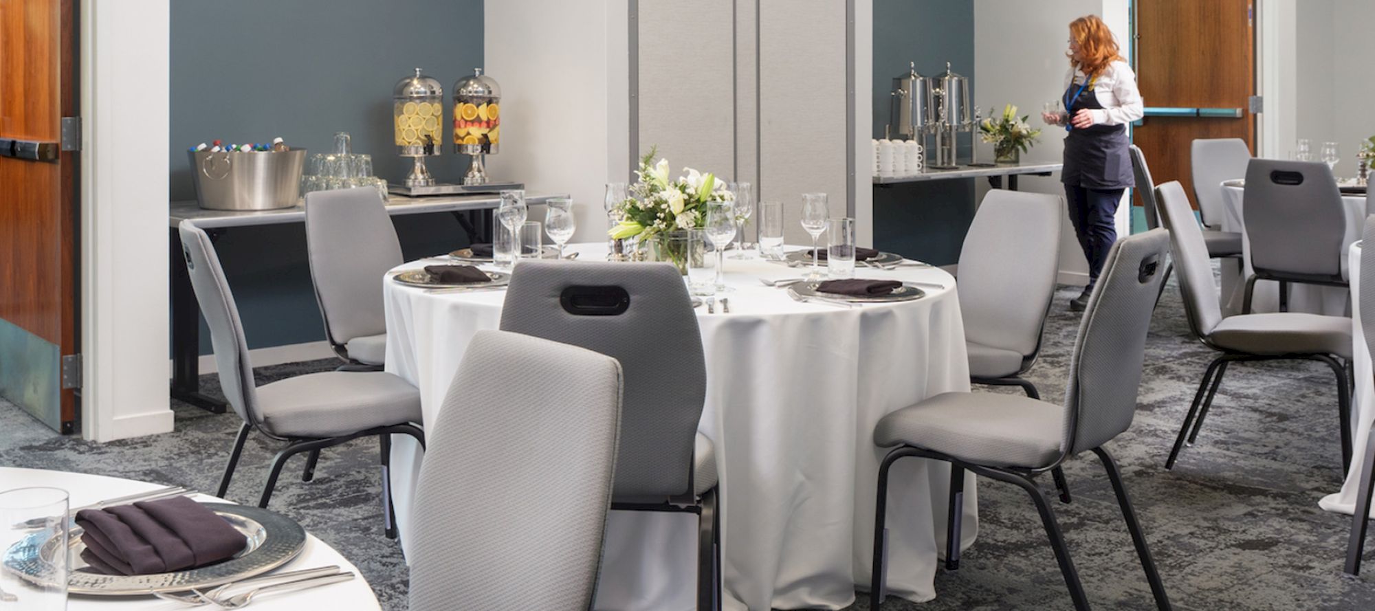 A banquet room set up with round tables, chairs, table settings, and beverages on a side table. A person is preparing the room.