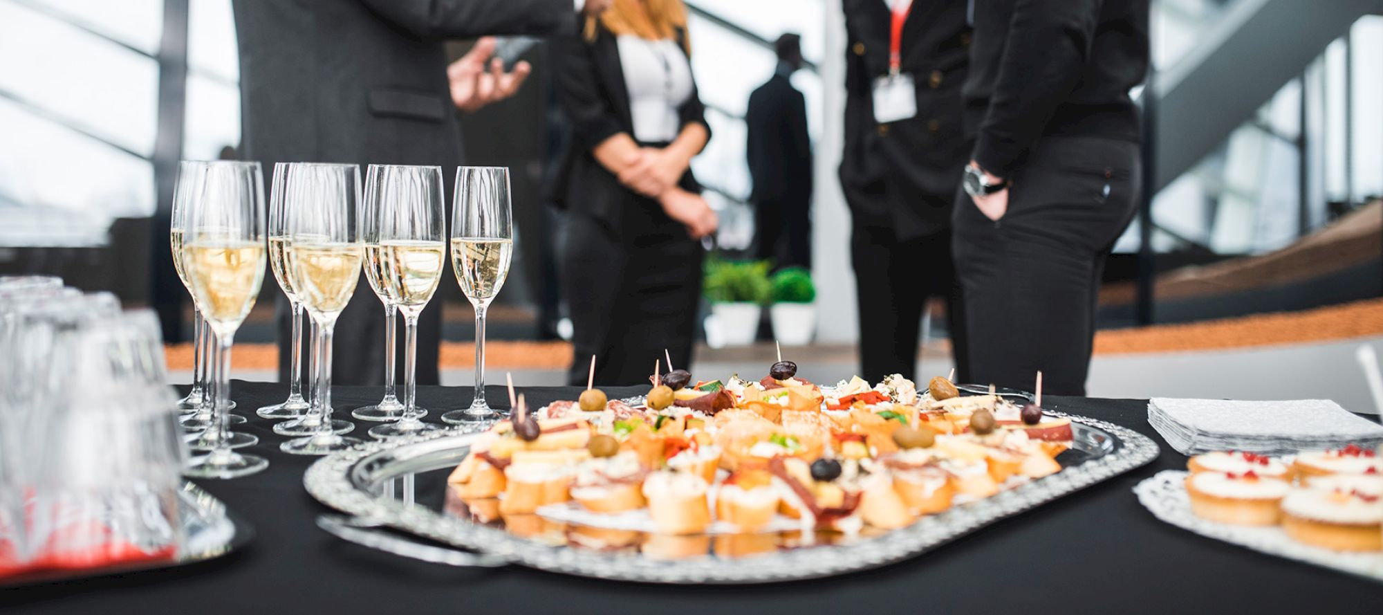 The image shows a table with champagne glasses and various appetizers, with people in formal attire conversing in the background.