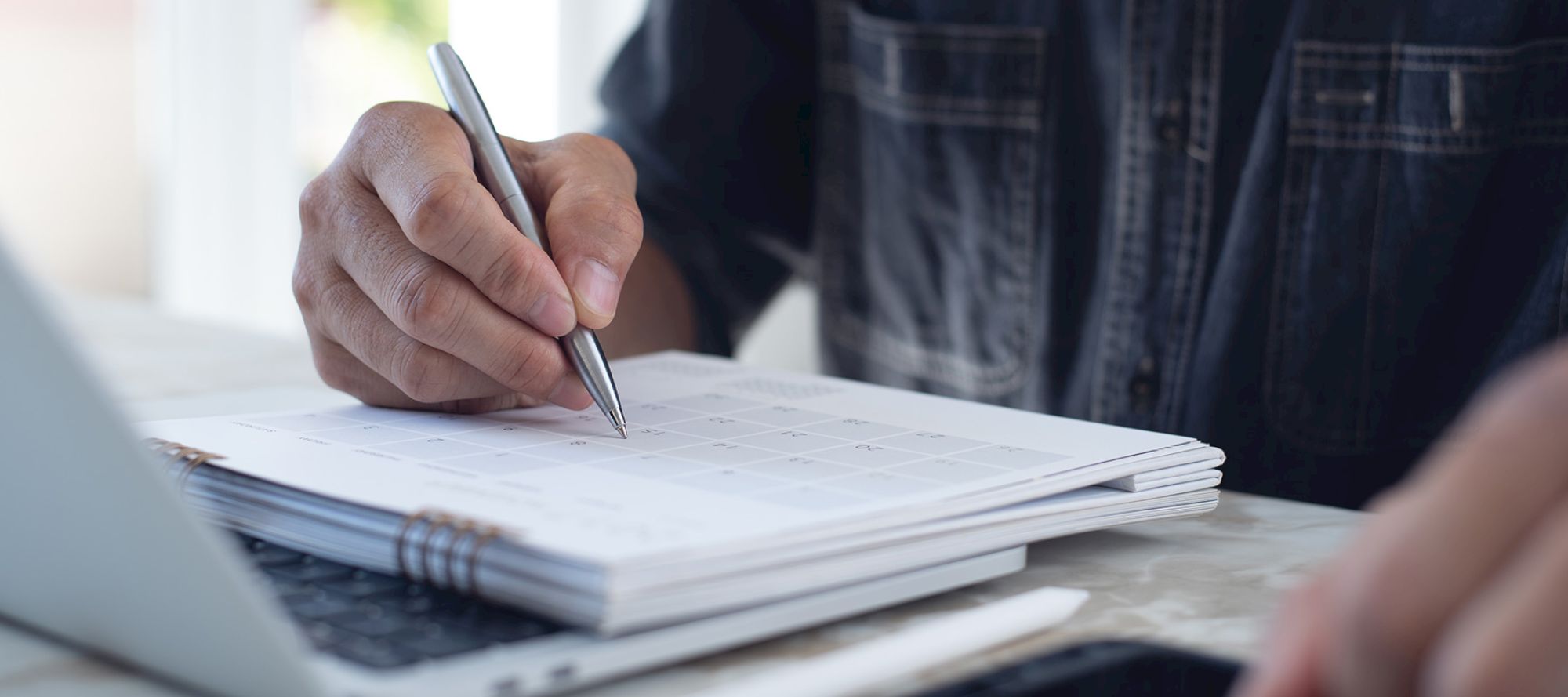 A person is writing on a calendar or planner with a pen, next to a laptop, likely organizing schedules or planning tasks for the day.