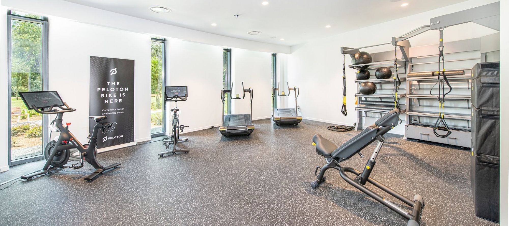 A modern gym features Peloton bikes, treadmills, weight equipment, and a functional training area with a bench, kettlebells, and TRX straps.