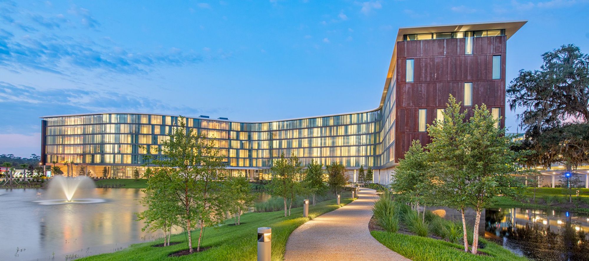 The image shows a modern, multi-story building with glass windows, a curved design, a water fountain, and a landscaped path with trees and lights ending the sentence.