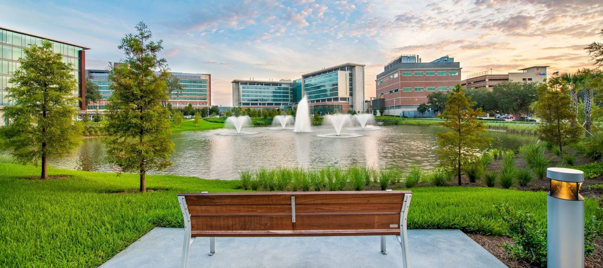 A wooden bench overlooks a pond with fountains, surrounded by greenery and modern buildings in the background, under a partly cloudy sky.