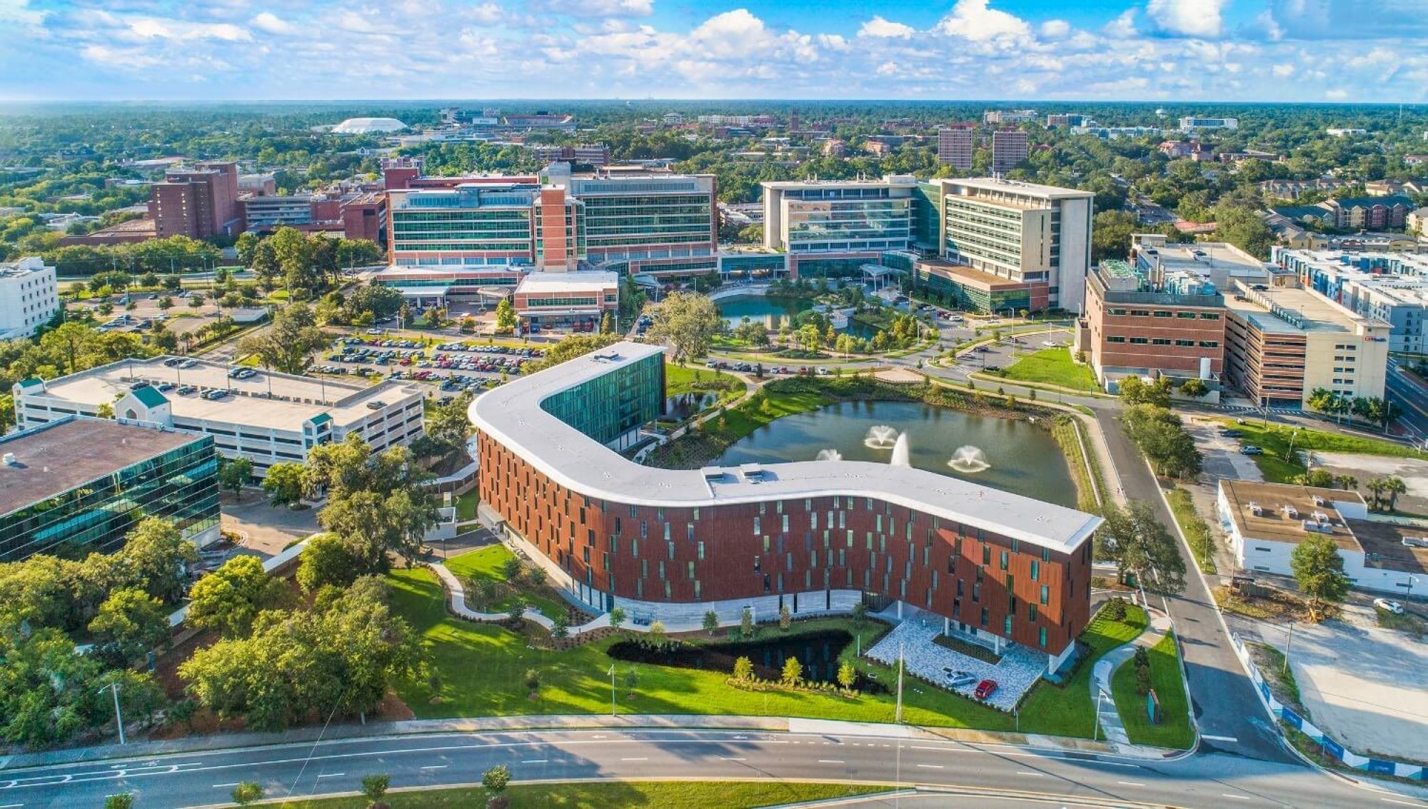 An aerial view of a modern campus with various buildings surrounded by greenery, a pond, and parking lots under a partly cloudy sky.