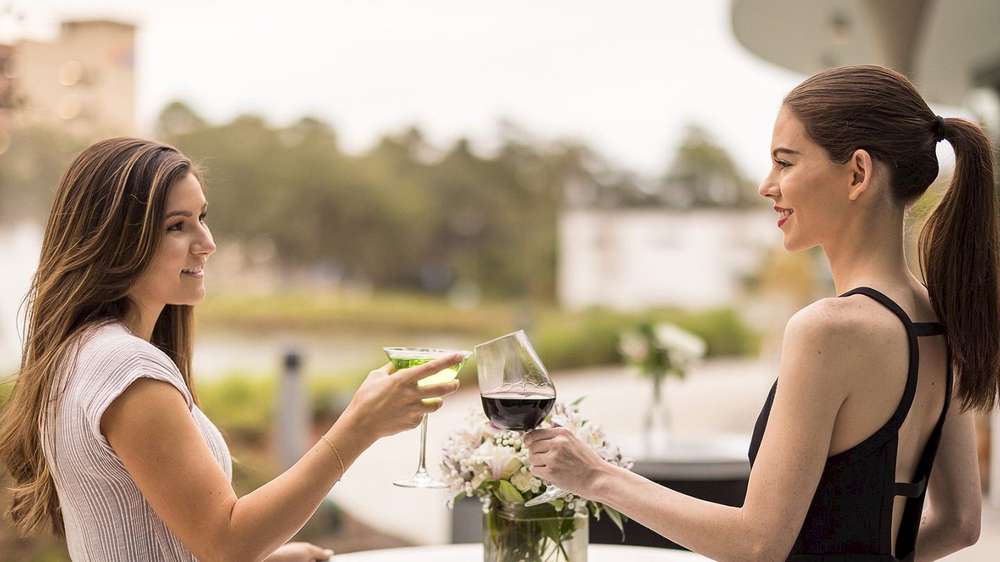 Two women clink glasses while standing outdoors; one holds a martini, and the other a glass of red wine, both smiling.