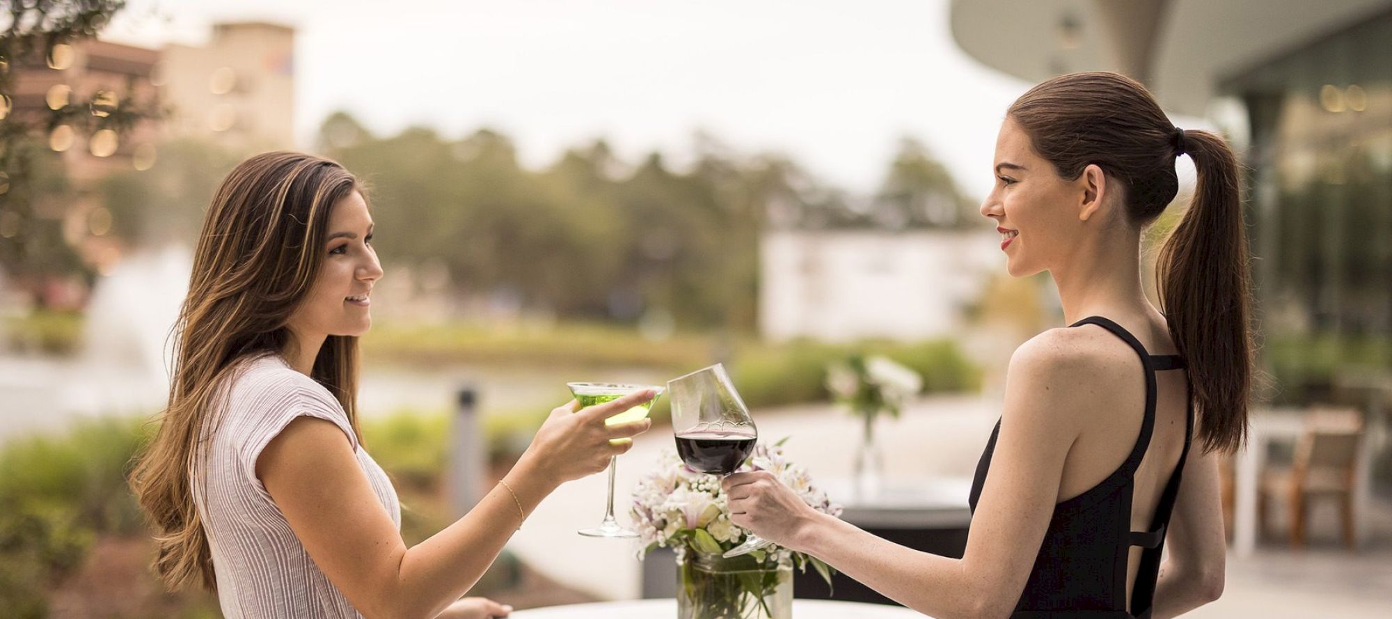 Two women clink glasses while standing outdoors; one holds a martini, and the other a glass of red wine, both smiling.