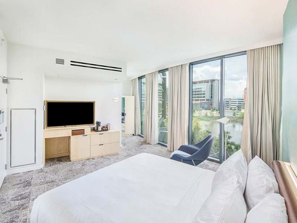 The image shows a modern hotel room with a large bed, a TV on a wooden cabinet, an armchair, and floor-to-ceiling windows with curtains.