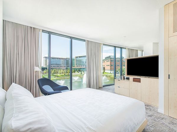 A modern bedroom with a large bed, flat-screen TV, blue chair, floor-to-ceiling windows, and a view of an urban landscape with buildings.