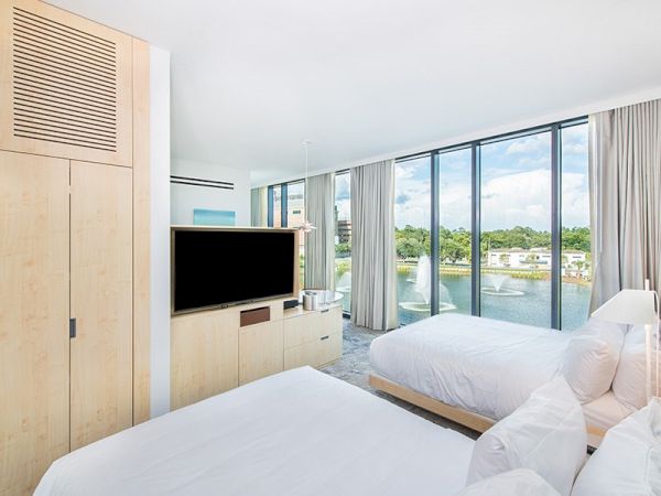 The image shows a modern hotel room with two beds, a large TV, wooden furniture, and large windows offering a scenic outdoor view.