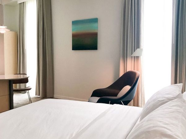 The image shows a modern, minimalistic bedroom with a neatly made bed, a cozy armchair, a framed painting, and natural light from a tall window.