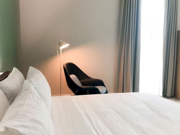 A modern bedroom featuring a neatly made bed with white sheets and pillows, a black chair, a standing lamp, and a window with light curtains.