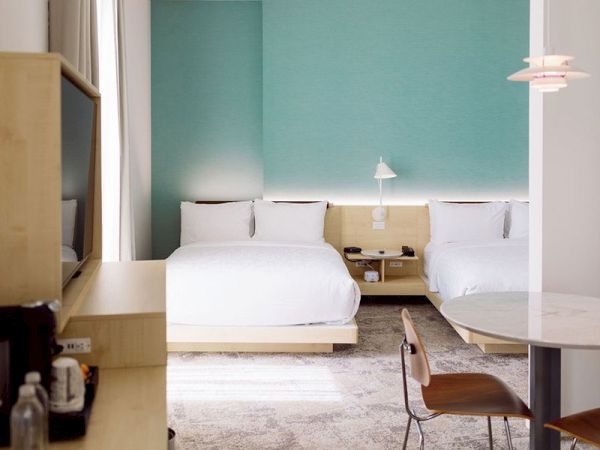 The image depicts a modern hotel room with two neatly made double beds, a TV, a desk, a chair, and a round table with another chair, ending the sentence.