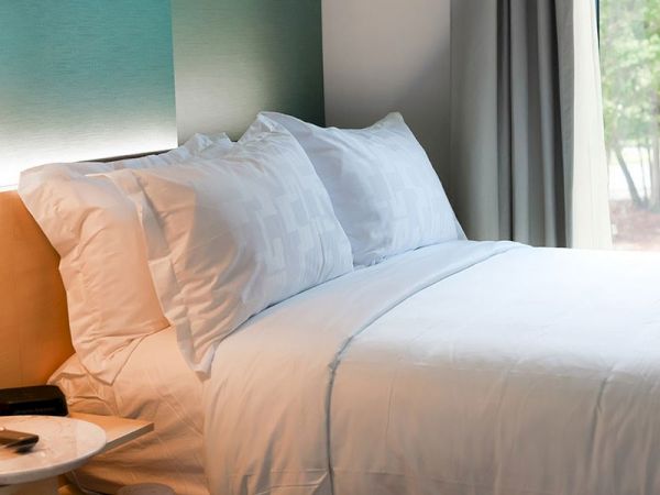 A neatly made bed with white sheets and pillows next to a small round table and a window. A TV remote is visible on the table.