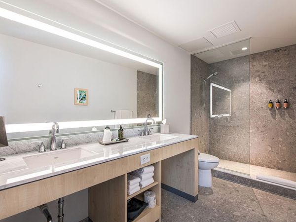 A modern bathroom featuring a double sink vanity with storage, a large backlit mirror, a toilet, and a spacious walk-in shower with dispensers.