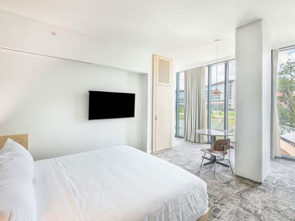 The image shows a modern, bright hotel room with a bed, mounted TV, small round table with two chairs, and large windows with a view outside.
