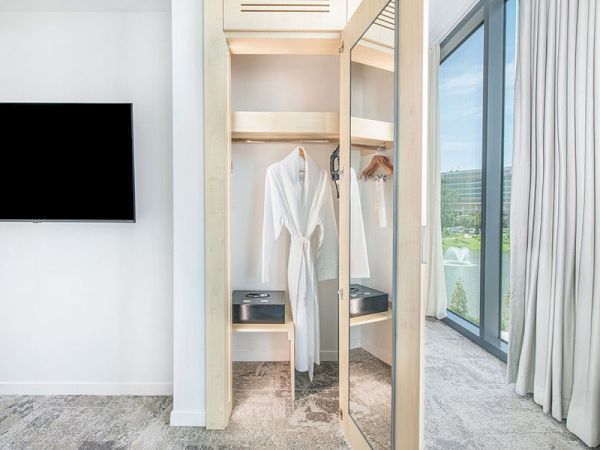 The image shows a modern hotel room with an open wardrobe holding a white bathrobe, a large mirror, a flat-screen TV, and a large window with a view.