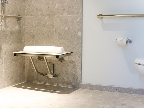 A bathroom with a folding shower seat, a towel, a toilet paper holder, and a grab bar, featuring tiled walls and floor.