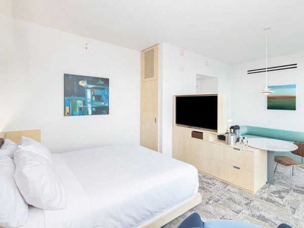 A modern hotel room features a bed, wall art, a TV, a coffee maker on a cabinet, and a seating area with a table and green bench.