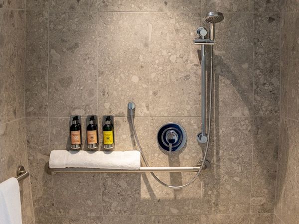 The image shows a tiled shower interior with a handheld showerhead, three bottles of toiletries on a towel rack, and a towel hanging on the side.