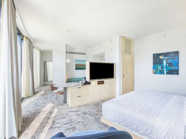 A modern hotel room with a bed, TV, desk, chairs, and large windows with curtains. The decor is minimalistic and bright, featuring abstract art.