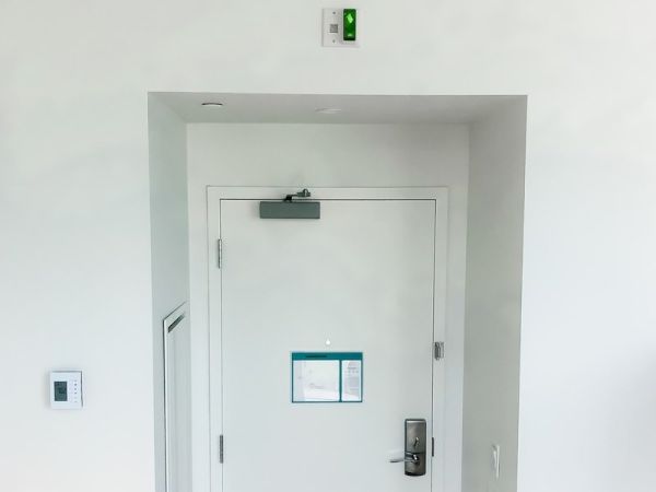 A white interior door with a push bar and an overhead closer is centered, flanked by walls, and topped with an emergency exit sign for guests with hearing impairments.