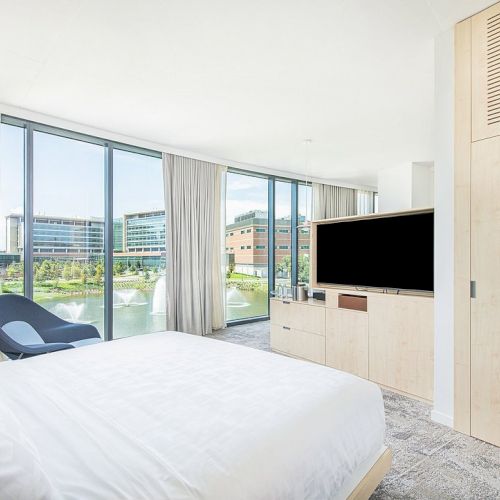 A modern bedroom with a double bed, TV, chair, and large windows overlooking buildings and a fountain, featuring light wooden furniture and neutral tones.