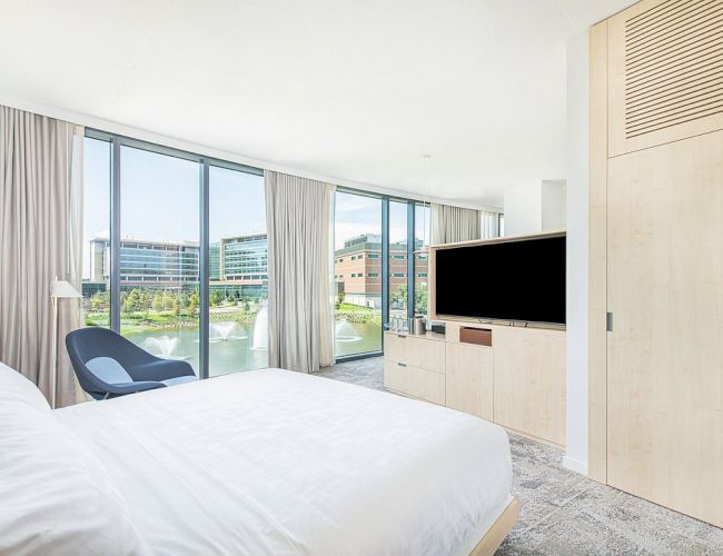 A modern bedroom with a double bed, TV, chair, and large windows overlooking buildings and a fountain, featuring light wooden furniture and neutral tones.