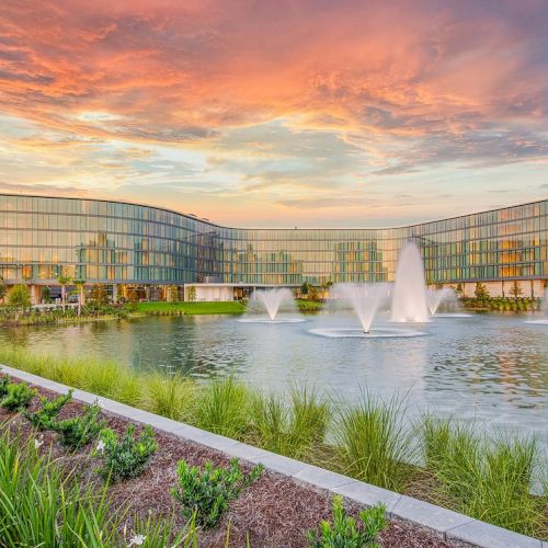 A modern building with glass windows stands behind a pond with three water fountains, under a vibrant, colorful sky during sunset.