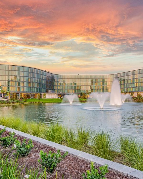 A modern building with glass windows stands behind a pond with three water fountains, under a vibrant, colorful sky during sunset.