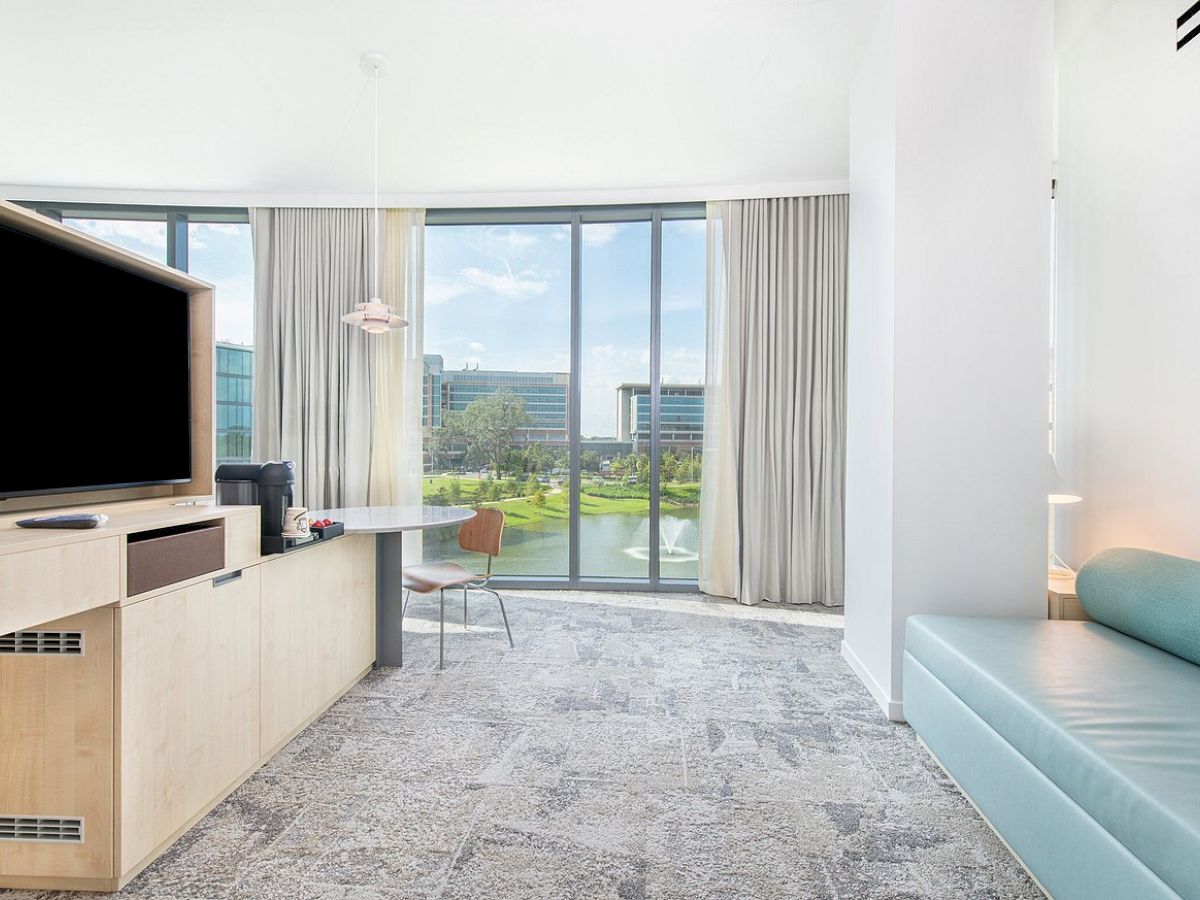 A modern hotel room with a TV, desk, chair, and couch. Large windows offer a view of a green park and buildings in the background.