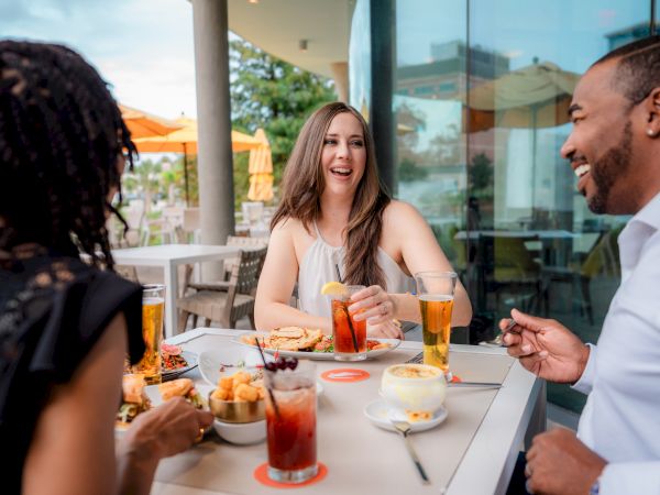 A group of friends cheers at an outdoor restaurant table, with a laughing woman holding a drink and enjoying a meal together.