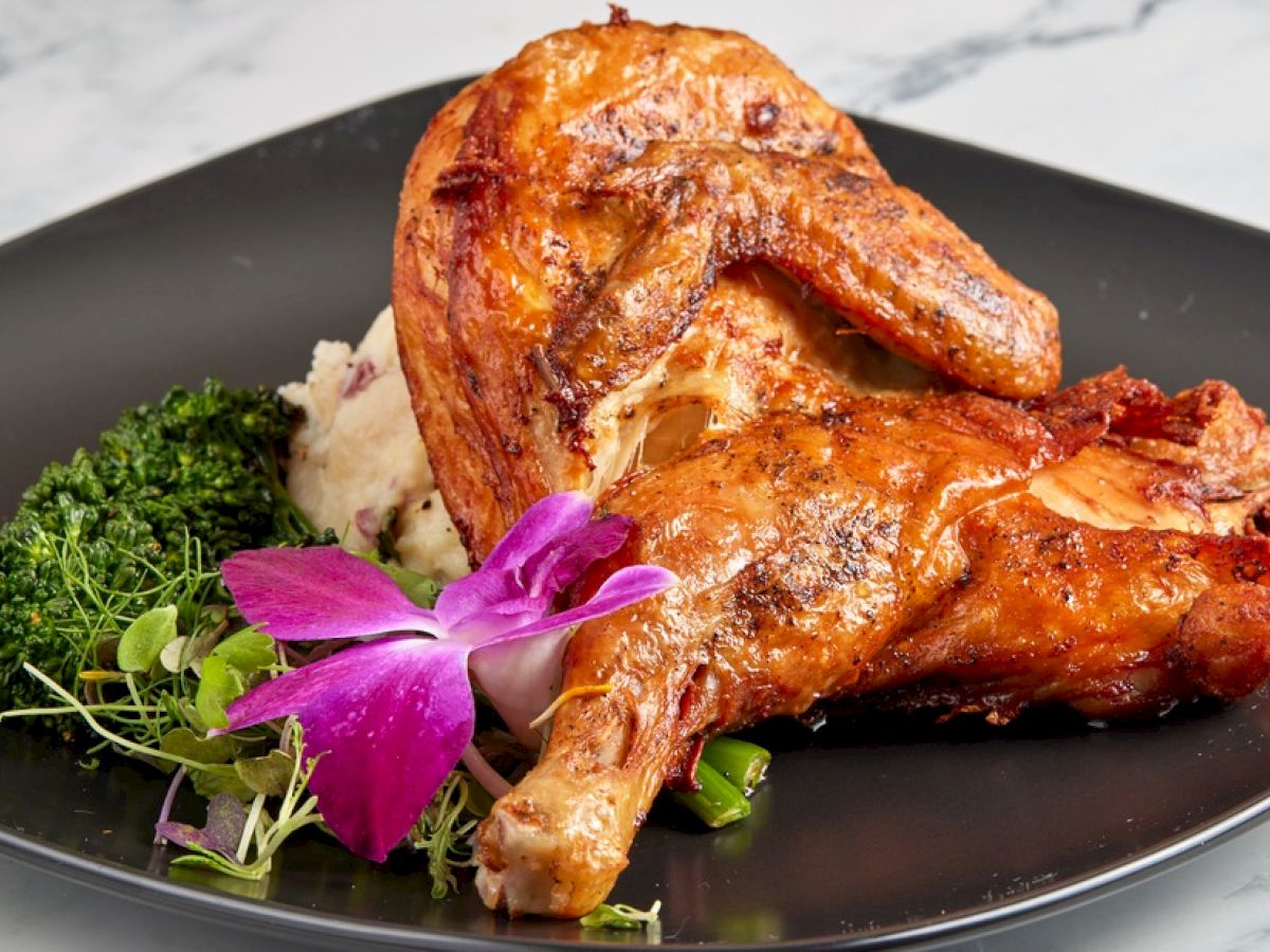 A plate with roasted chicken, mashed potatoes, and broccoli garnished with a purple flower on a dark plate.