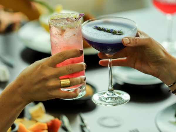 Two people are clinking glasses, one holding a pink cocktail and the other a blue cocktail, with dishes of food on the table.