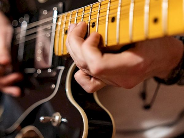 A close-up shot of a person playing a black electric guitar, focusing on their hands and the guitar fretboard, with a blurred background.