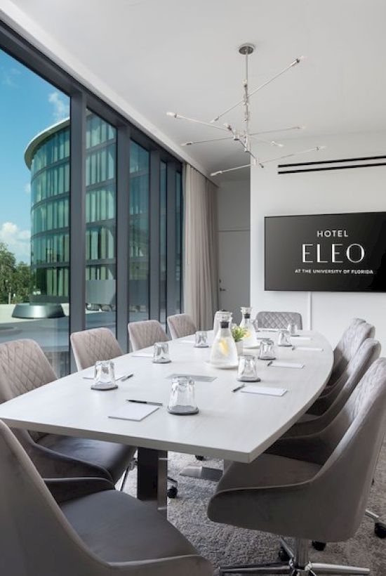 A modern conference room with a long table, grey chairs, and a large window showing an outside view; "Hotel Eleo" is displayed on a screen on the wall.