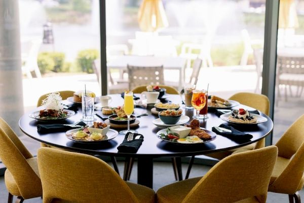 A round table set for six with various dishes, drinks, and yellow chairs near large windows overlooking a patio with fountains and greenery.