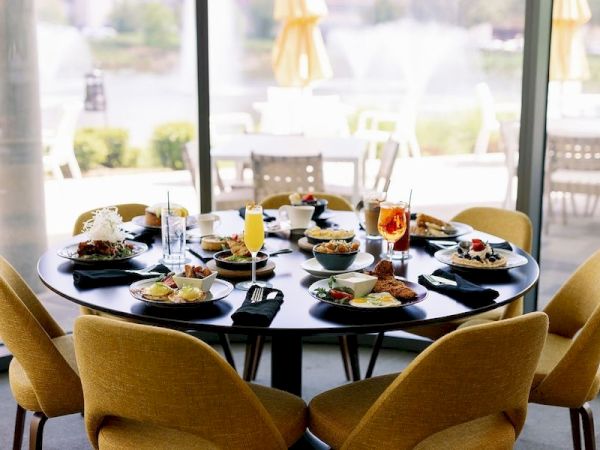 A round table set for six with various dishes, drinks, and yellow chairs near large windows overlooking a patio with fountains and greenery.