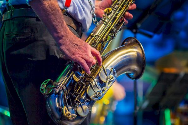 A close-up of a person in a blue shirt and red suspenders playing a saxophone on stage, with blurred blue and yellow lights in the background.