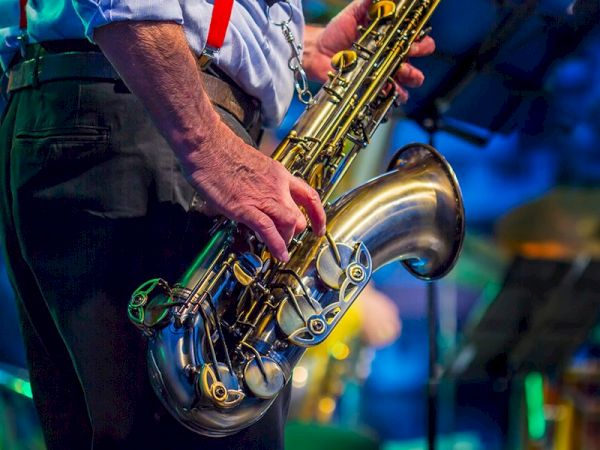 A close-up of a person in a blue shirt and red suspenders playing a saxophone on stage, with blurred blue and yellow lights in the background.