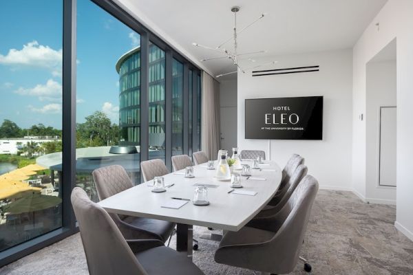 The image shows a modern conference room with a long table, chairs, a TV screen displaying 