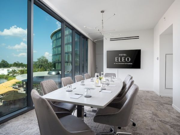 The image shows a modern conference room with a long table, chairs, a TV screen displaying "Hotel Eleo," and large windows offering an exterior view.