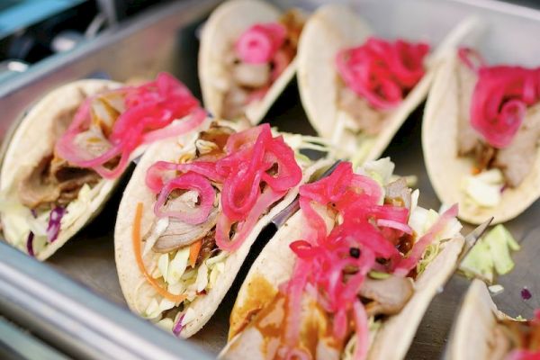 The image shows a tray filled with tacos, each topped with what appears to be pickled red onions and cabbage, along with other ingredients.