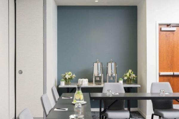 The image shows a conference room setup with tables and chairs. There's a refreshment area with beverage dispensers and flowers on a table in the back.