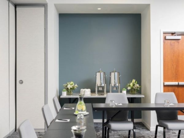 The image shows a conference room setup with tables and chairs. There's a refreshment area with beverage dispensers and flowers on a table in the back.