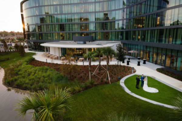 A modern glass building with a grassy landscape features a wedding photoshoot, with a couple posing next to the water.