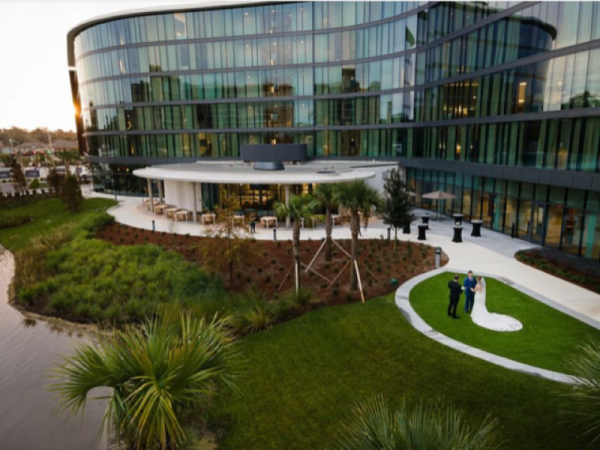 A modern glass building with a grassy landscape features a wedding photoshoot, with a couple posing next to the water.